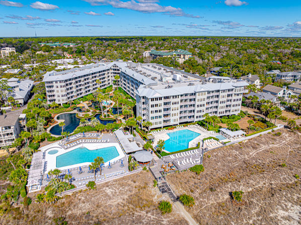 Aerial view of a sunny beachfront resort complex with multi-story oceanfront condos, two large pool decks, winding landscaped lagoons, palm trees and a boardwalk crossing coastal dunes to the beach — bright coastal vacation scene