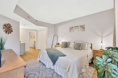 Airy master bedroom with king bed in neutral linens, green throw and accent pillows, wood nightstands with pendant lamps, full-length mirror, patterned rug, hardwood floors, visible en-suite bathroom and a large potted plant.