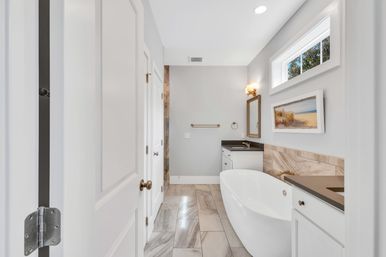 Bright spa-like modern bathroom with freestanding oval tub, gray marble floor tiles, white vanities with dark countertops, brass fixtures, coastal wall art, and a small horizontal window.