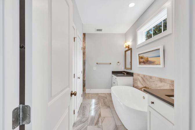 Bright spa-like modern bathroom with freestanding oval tub, gray marble floor tiles, white vanities with dark countertops, brass fixtures, coastal wall art, and a small horizontal window.