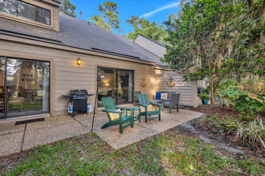 Backyard patio with green Adirondack chairs and a bench with nautical cushions, gas grill by sliding glass doors of a wood-sided house, concrete patio surrounded by trees draped in Spanish moss