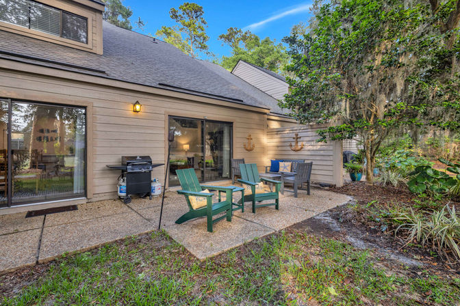 Backyard patio with green Adirondack chairs and a bench with nautical cushions, gas grill by sliding glass doors of a wood-sided house, concrete patio surrounded by trees draped in Spanish moss