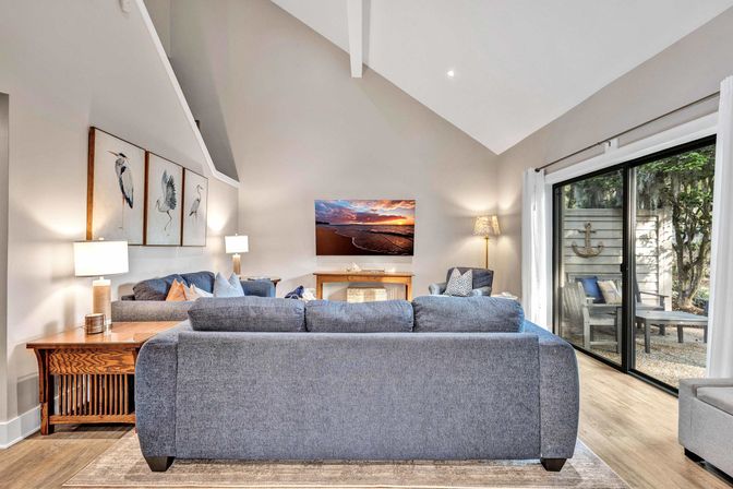 Cozy modern coastal living room with vaulted ceiling and blue-gray sofas facing a wall-mounted TV over a wooden console, bird artwork, warm table lamps, and sliding glass doors opening to a patio with outdoor seating and decorative anchor.