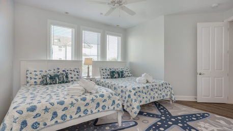 Bright coastal vacation rental bedroom with two matching beds dressed in blue shell-and-starfish quilts, folded towels on each bed, bedside lamp between three windows with blinds, ceiling fan, and starfish rug.