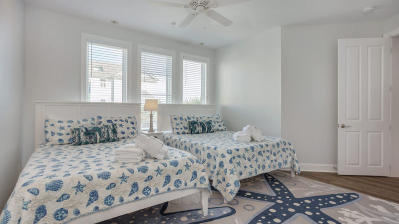 Bright coastal vacation rental bedroom with two matching beds dressed in blue shell-and-starfish quilts, folded towels on each bed, bedside lamp between three windows with blinds, ceiling fan, and starfish rug.