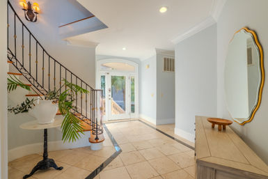 Sunlit coastal-style foyer with marble tile floor, curved wooden staircase with wrought-iron railing, palm-frond arrangement on a pedestal, and a wooden console under an oval mirror.