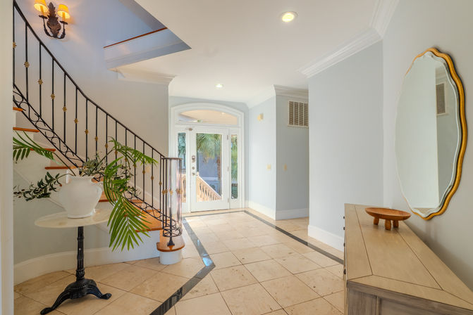 Sunlit coastal-style foyer with marble tile floor, curved wooden staircase with wrought-iron railing, palm-frond arrangement on a pedestal, and a wooden console under an oval mirror.