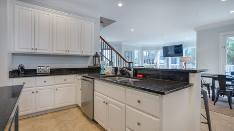 Bright open-concept kitchen with white cabinets, black granite countertops, stainless dishwasher and undermount sink, breakfast bar overlooking living and dining area with staircase, large windows, and recessed lighting.
