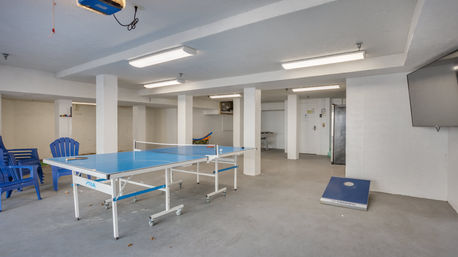 Bright finished basement game room with blue ping pong table on casters, cornhole board, blue plastic chairs, wall-mounted TV, white support columns and fluorescent ceiling lights on a concrete floor.