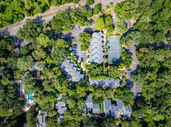 Aerial bird's-eye view of a tree‑canopied residential complex with gray-roofed townhomes, tennis court, swimming pool and parking areas