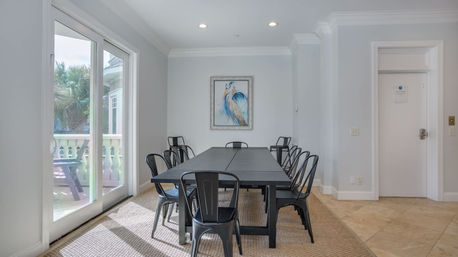 Sunlit coastal dining room with a long black table and eight black metal chairs, sliding glass doors to a palm-lined balcony and framed heron artwork on a pale gray wall.