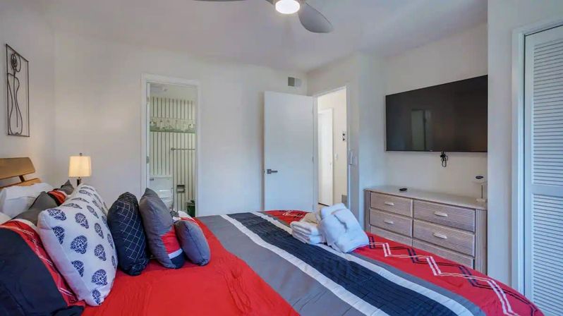 Bright modern bedroom with red, gray, and navy striped bedding, layered decorative pillows and folded white towels on the bed, wall-mounted flat-screen TV above a light-wood dresser, ceiling fan, and open door revealing an en-suite bathroom.