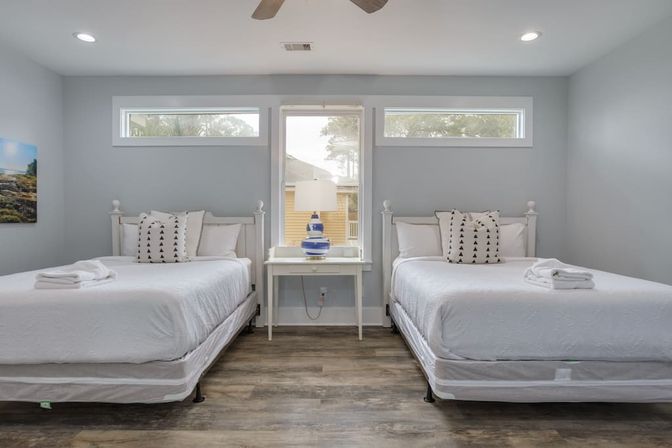 Airy coastal-style guest bedroom with two matching white beds, patterned throw pillows and folded towels, blue-and-white lamp on a central nightstand, transom windows and wood-look floors.