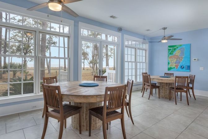 Airy coastal dining room with light-blue walls, two round wooden tables and chairs, tile floor, ceiling fans and large windows overlooking a marsh and distant water.