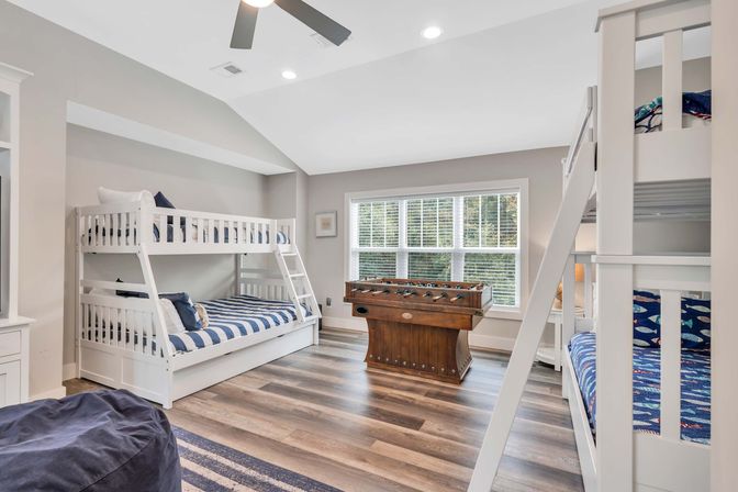Bright coastal-style kids' bedroom with white bunk beds, blue striped bedding, hardwood floors, a large window with blinds, and a wooden foosball table