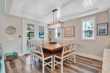 Bright coastal dining room in a beach house: wooden farmhouse table with six white chairs, geometric pendant light, French doors to a patio, “Beach People Welcome” wall sign, shiplap ceiling and wide-plank wood floors.