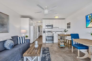 Bright coastal-style condo interior with white kitchen and stainless-steel appliances, navy sofa with striped pillows, wooden coffee table, blue dining chairs and ceiling fan.
