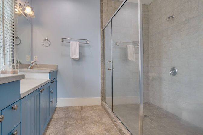 Bright modern bathroom with blue beadboard vanity and quartz countertop, glass-enclosed tiled walk-in shower, beige tile floor, light blue walls, chrome fixtures and a white towel.