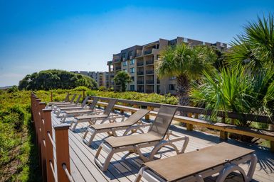 Sunny coastal boardwalk lined with reclining lounge chairs, palm trees and mid-rise beachfront condos under a clear blue sky