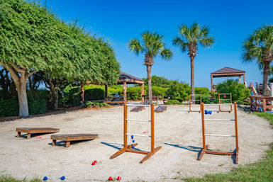 Sunny beachside recreation area with palm trees and picnic shelters, sandy courts set up for cornhole and ladder toss lawn games under a clear blue sky.