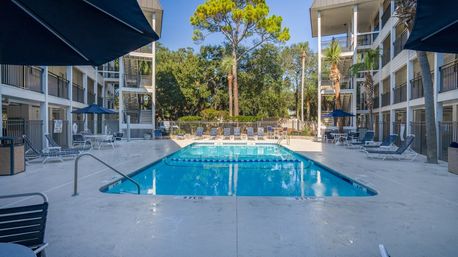 Sunny outdoor hotel courtyard pool between two balcony-lined buildings, lounge chairs and blue umbrellas, palm trees and tall pines in the background.