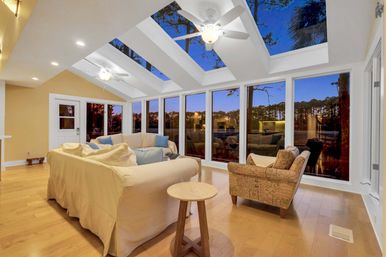 Cozy sunroom with vaulted skylights and ceiling fans, beige slipcovered sofa and patterned armchair on hardwood floors, floor-to-ceiling windows framing a twilight pine-lined lake.