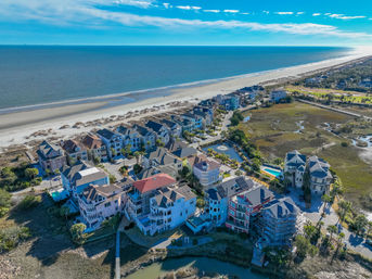 Aerial shot of colorful oceanfront beach houses perched along a wide sandy shoreline, with marshy wetlands, backyard pools and the sparkling blue ocean stretching to the horizon.