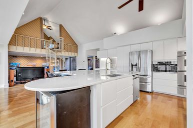 Airy modern open-concept kitchen in a loft-style home — curved white quartz island with sink, stainless-steel fridge and ovens, sleek white cabinetry, warm hardwood floors, vaulted ceiling and spiral staircase up to a wood-paneled loft.