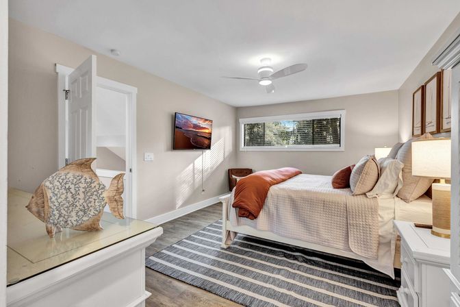 Bright contemporary coastal bedroom with neutral walls and white trim, ceiling fan, wall-mounted TV, large window with blinds, striped rug, decorative wooden fish sculpture on a dresser, and a neatly made bed with a rust-colored throw.