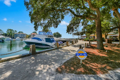 Sunny coastal marina with a docked white yacht along an oak‑shaded waterfront promenade, benches, a small tour sign and brick walkway in a charming coastal town