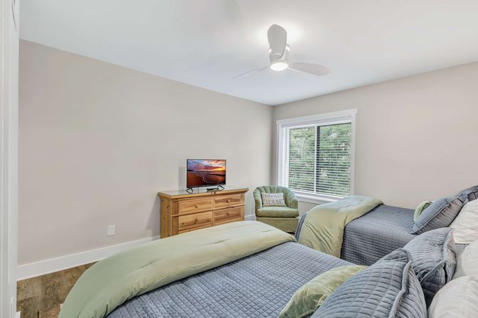 Cozy coastal-style bedroom with two twin beds in blue-gray quilts and green duvets, wooden dresser with TV, green armchair by a window with blinds, white ceiling fan and neutral walls
