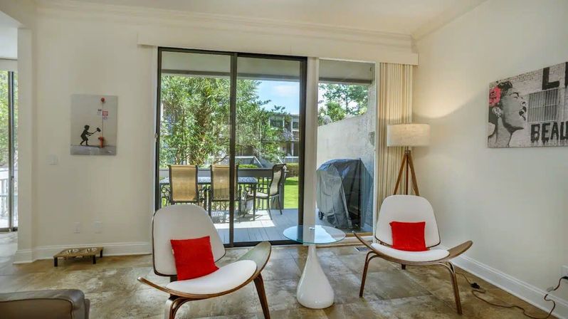 Bright modern living room with two white mid-century chairs and red accent pillows, glass-top side table and tripod floor lamp, sliding glass doors opening to a patio with outdoor dining set, grill and garden view.