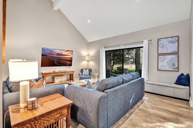 Cozy sunlit living room with vaulted ceiling in a suburban home featuring a gray sectional sofa, matching armchair, wooden side table and console, wall-mounted TV showing a sunset beach, sliding glass doors to a tree-lined patio, hardwood floors and neutral artwork.