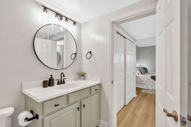 Bright modern ensuite bathroom with round wall mirror, white quartz countertop vanity with bronze faucet and muted green cabinets, exposed-bulb vanity light, toilet, and open door revealing bedroom with wood floors and white sliding closet doors.