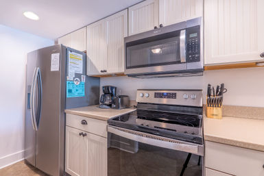 Bright modern kitchen corner with stainless-steel refrigerator, over-the-range microwave, electric glass-top stove, white paneled cabinets, beige countertops, coffee maker, toaster, and knife block