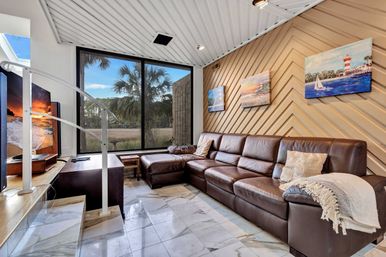 Cozy coastal living room with a large brown leather sectional, chevron wood accent wall displaying lighthouse paintings, marble tile floor, spiral staircase, and a floor-to-ceiling window framing palm trees and a marsh view.