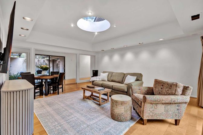 Sunlit open-concept living room with round skylight and recessed lighting, hardwood floors, neutral sofa, patterned armchair and ottoman, wooden coffee table on a pale geometric rug, and a dining nook by sliding glass doors to a patio.