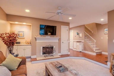 Cozy beige living room interior with wall-mounted TV above a white mantel, decorative fireplace screen, ceiling fan and recessed lighting, brown sofa, coffee table, hardwood entry and carpeted sunken seating area with staircase.