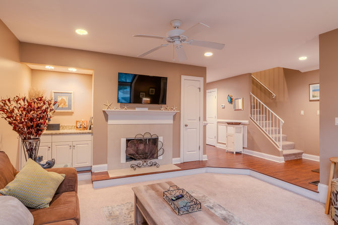 Cozy beige living room interior with wall-mounted TV above a white mantel, decorative fireplace screen, ceiling fan and recessed lighting, brown sofa, coffee table, hardwood entry and carpeted sunken seating area with staircase.