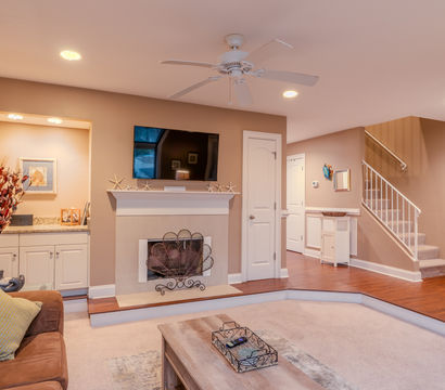 Cozy beige living room interior with wall-mounted TV above a white mantel, decorative fireplace screen, ceiling fan and recessed lighting, brown sofa, coffee table, hardwood entry and carpeted sunken seating area with staircase.