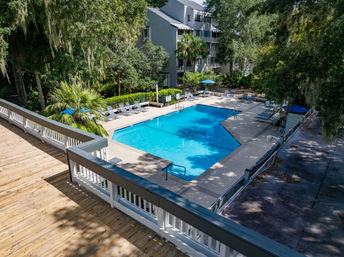 Aerial view of a sunlit outdoor pool at a condo complex: bright blue water, lounge chairs and umbrellas on a concrete deck, surrounded by palm trees and oaks draped in Spanish moss, wooden deck railing in the foreground.
