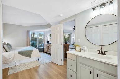 Bright master bedroom with en-suite vanity and round mirror, king bed with neutral bedding, seating area and sliding glass doors to a balcony framed by palm trees, light wood floors.