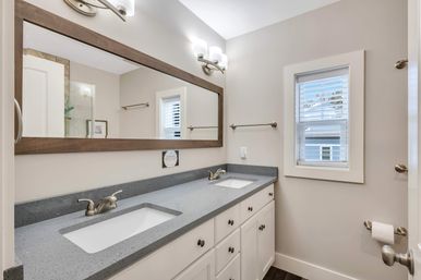 Bright residential bathroom with double sinks on a gray quartz countertop, white cabinets, large wood-framed mirror, brushed-nickel faucets, wall sconces and a window with blinds.