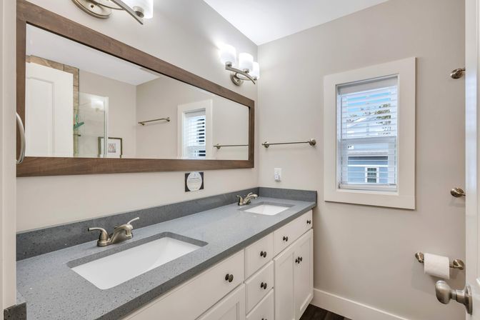 Bright residential bathroom with double sinks on a gray quartz countertop, white cabinets, large wood-framed mirror, brushed-nickel faucets, wall sconces and a window with blinds.