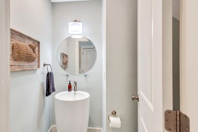 Cozy modern powder room in a home featuring a white cylindrical pedestal sink, round mirror with glass shelf, overhead sconce, amber soap dispenser, towel ring, woven wall art and toilet paper holder.