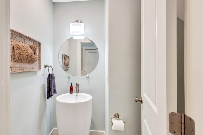 Cozy modern powder room in a home featuring a white cylindrical pedestal sink, round mirror with glass shelf, overhead sconce, amber soap dispenser, towel ring, woven wall art and toilet paper holder.