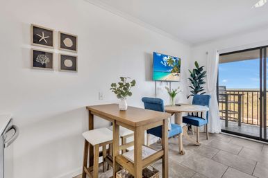 Bright coastal dining area in a beachfront condo: wooden bar table with stools, round table with blue upholstered chairs, potted plants, wall TV showing a tropical beach, and sliding glass doors to a balcony ocean view.