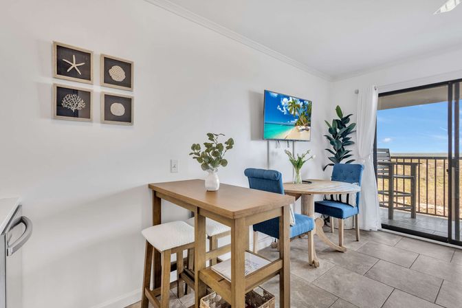 Bright coastal dining area in a beachfront condo: wooden bar table with stools, round table with blue upholstered chairs, potted plants, wall TV showing a tropical beach, and sliding glass doors to a balcony ocean view.