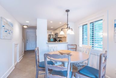 Sunlit coastal-inspired dining area with round wooden table, blue-cushioned ladder-back chairs, wicker centerpiece, hanging pendant light, white plantation shutters and an open kitchen with stainless steel appliances
