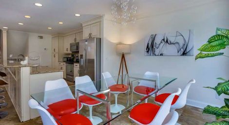 Contemporary open-plan kitchen and dining area with a glass table, white chairs with red cushions, marble island, stainless-steel appliances, abstract wall art and tripod floor lamp.